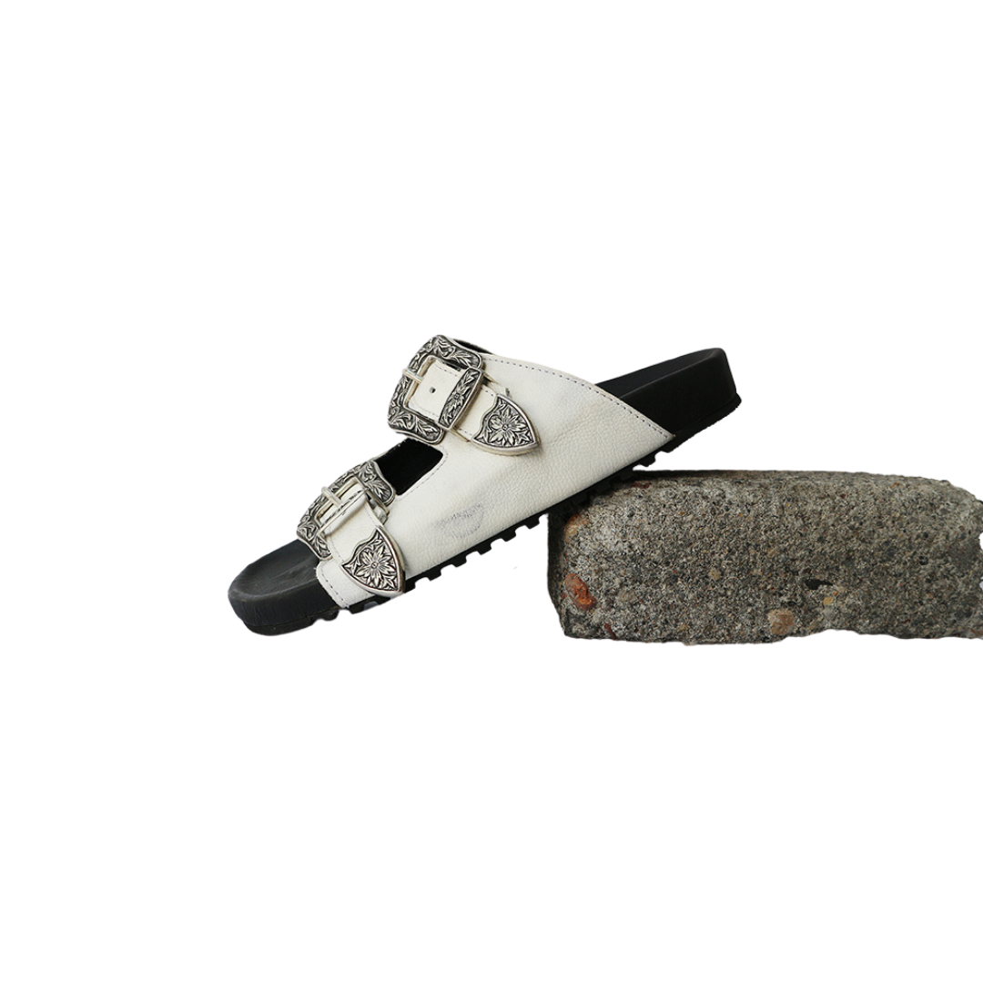 The image of a white sandal made of cowhide, designed with two straps and a carved buckle decoration is the Becky Slides Sandal from Seminyak Leather Bali