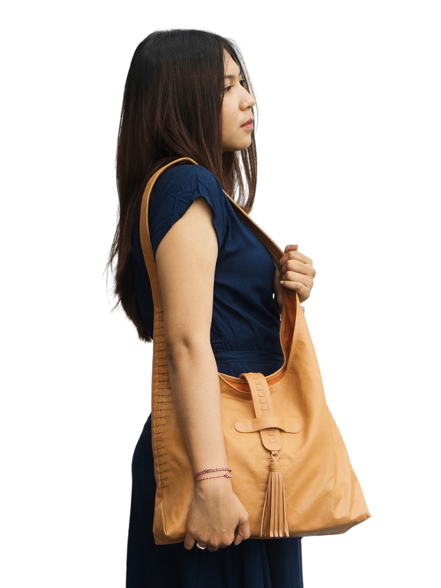 The picture shows a model with a blue dress holding a honey tan bag. The bag is made from goat leather, with a woven detail along the bag and a tassel detail in front on of the magnetic closure. The bag dimension is 41 x 31 x 10 cm. It has 1 main compartment and 1 lining pocket also a phone pocket on it. It is Ella Hobo Bag in Honey Tan from Seminyak Leather Bali.