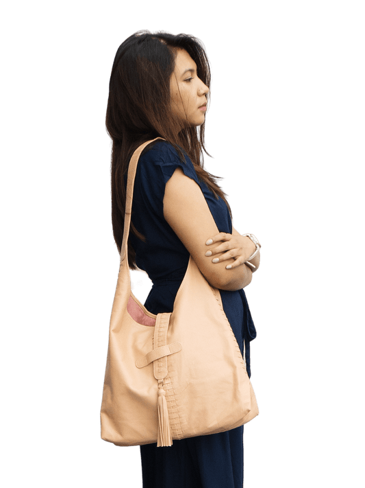 A picture shows a model in long blue dress with a bag hanging in her shoulder. The bag has a woven detail crossed in side to side and front to back, with a leather tassel in the magnetic closure. It is Ella Hobo Bag in Nude Pink from Seminyak Leather Bali.