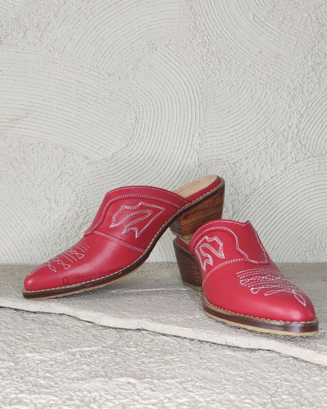 Pair of red pointed-toe mules with white embroidery designs by Seminyak Leather Bali on a light gray background