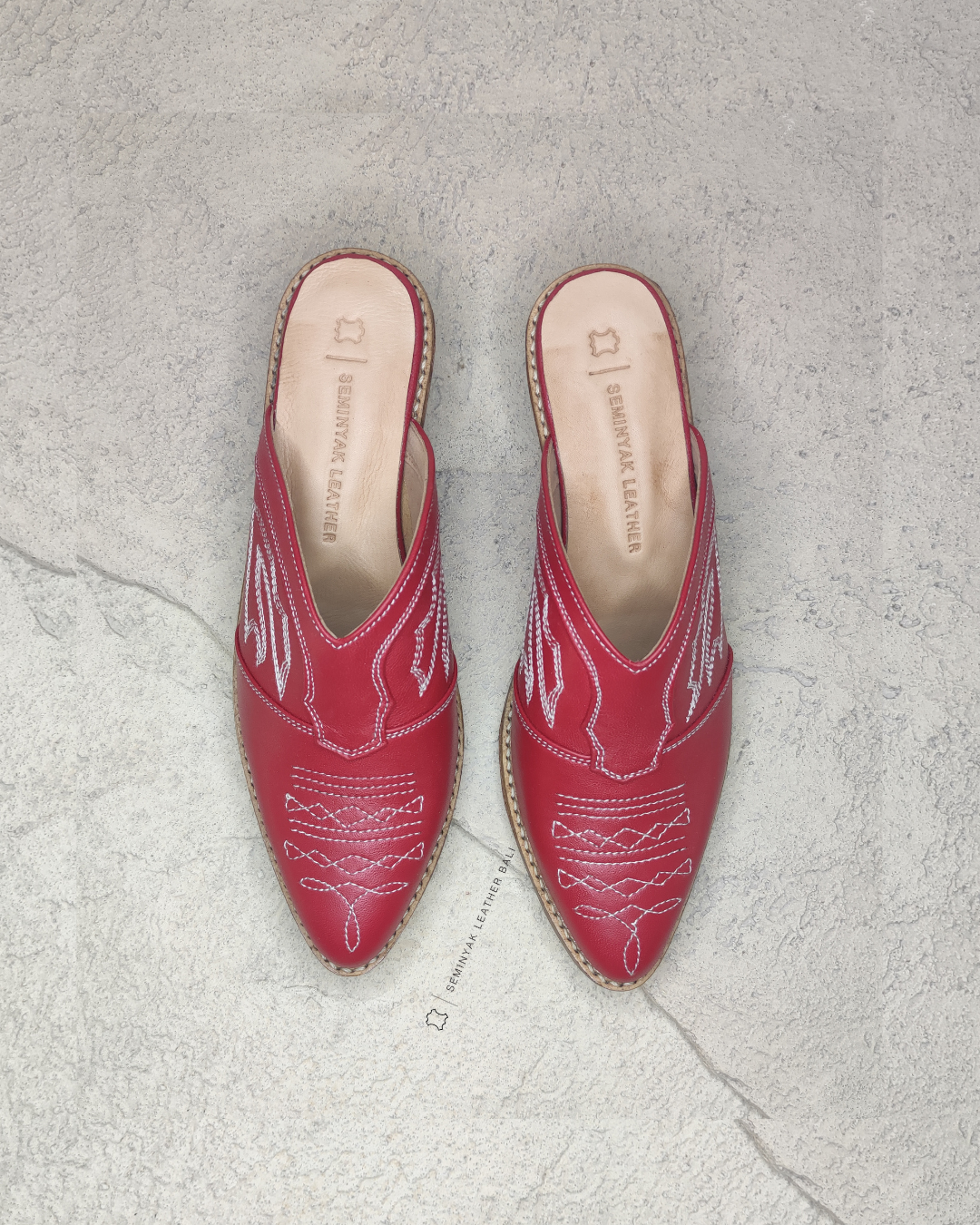 Pair of red pointed-toe mules with white embroidery designs by Seminyak Leather Bali on a light gray background