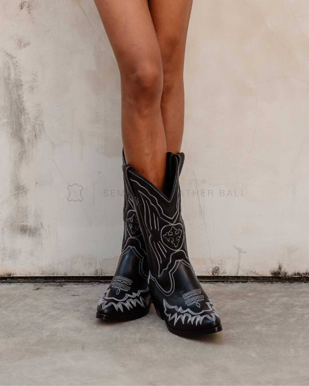 Black cowboy boots with white embroidery worn by a person designs by Seminyak Leather Bali on a neutral background