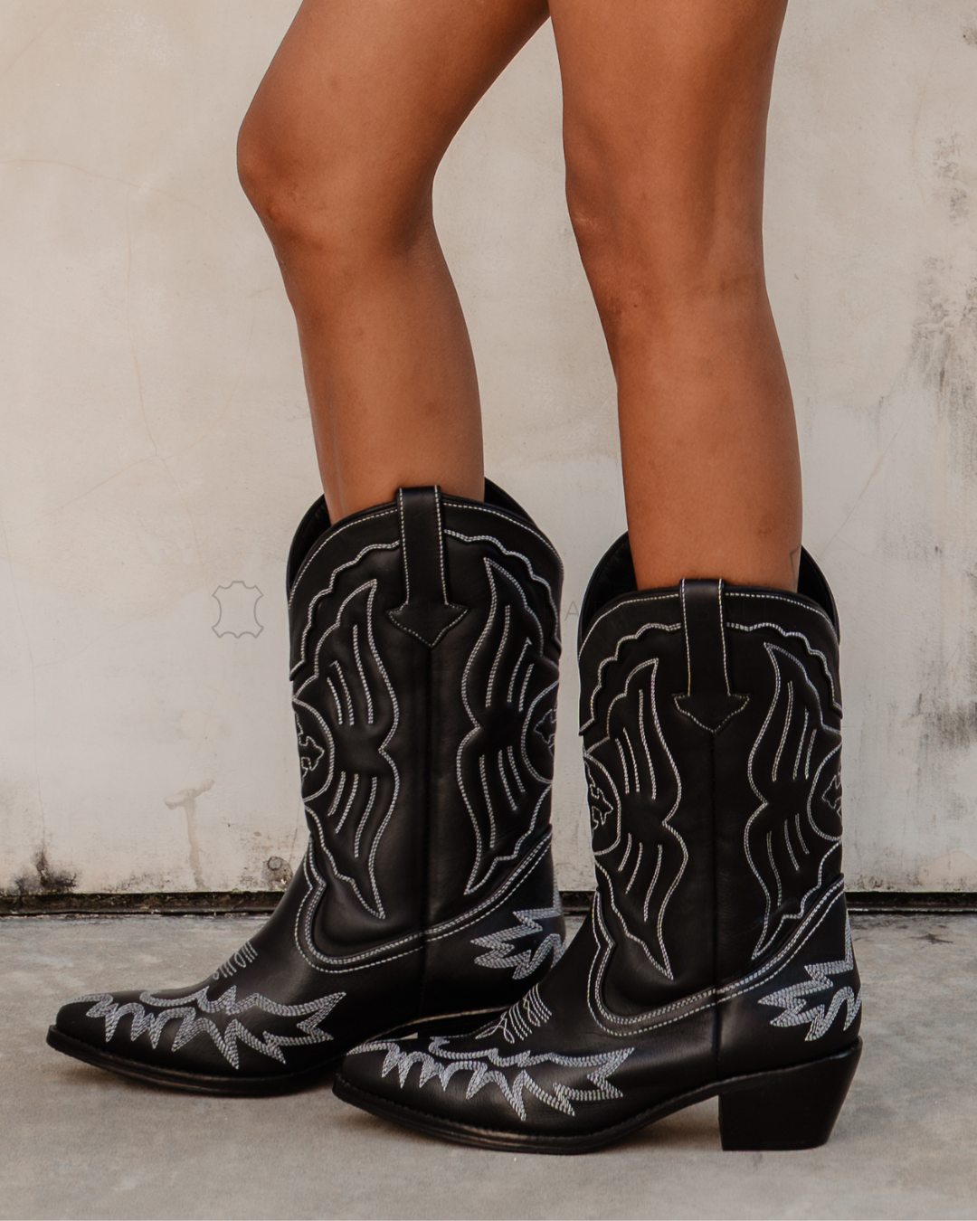 Black cowboy boots with white embroidery worn by a person designs by Seminyak Leather Bali on a neutral background