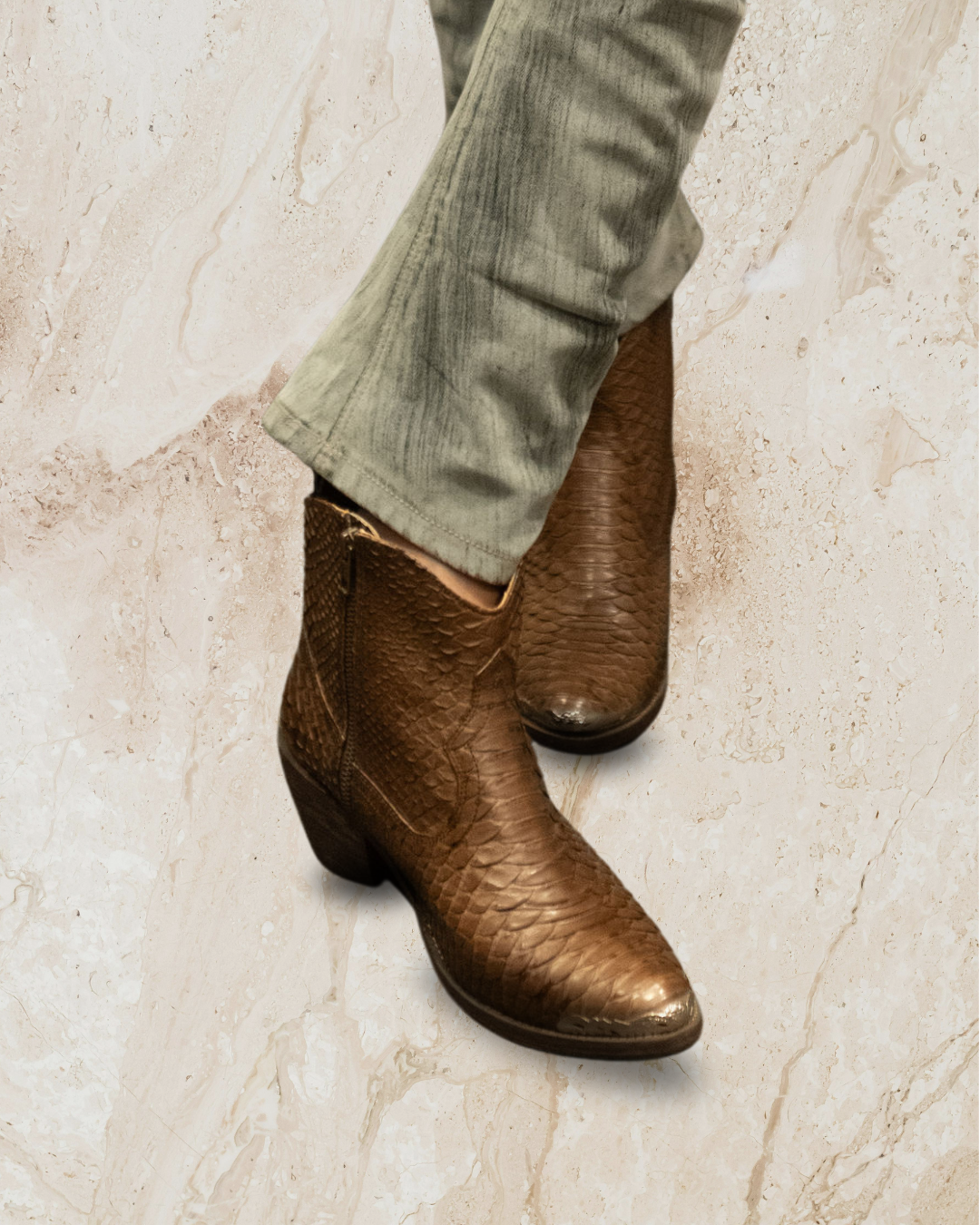 Snake skin ankle boots worn with light-colored pants designs by Seminyak Leather Bali on a textured beige background