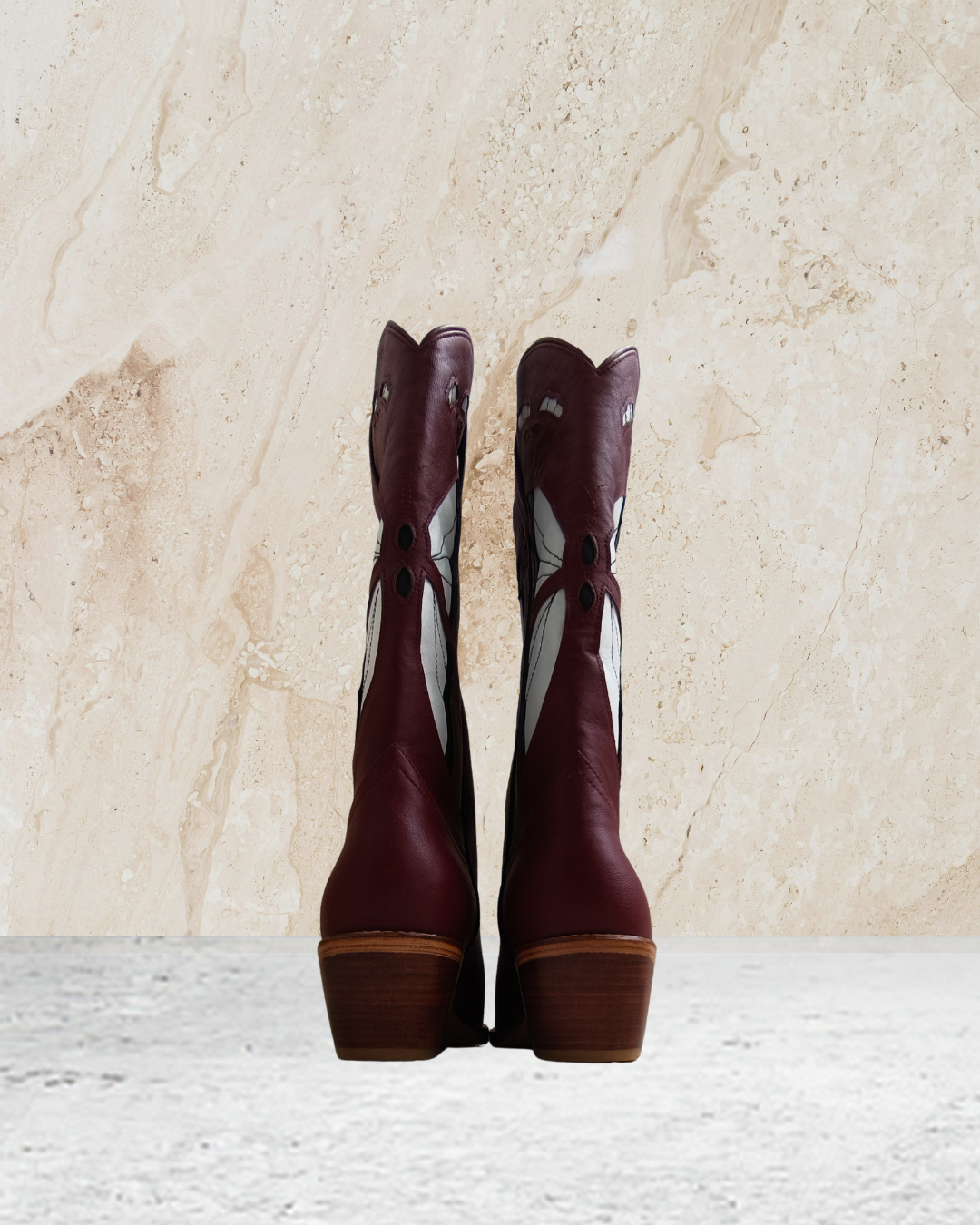 Pair of maroon cowboy boots with butterfly designs by Seminyak Leather Bali on a textured beige background