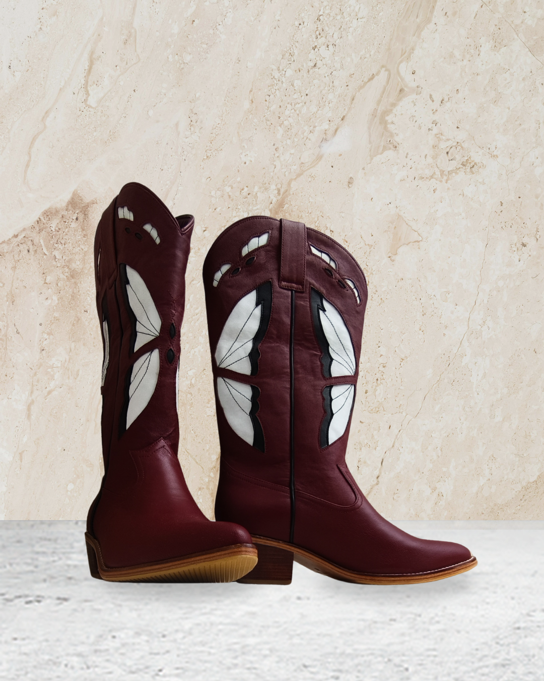 Pair of maroon cowboy boots with butterfly designs by Seminyak Leather Bali on a textured beige background