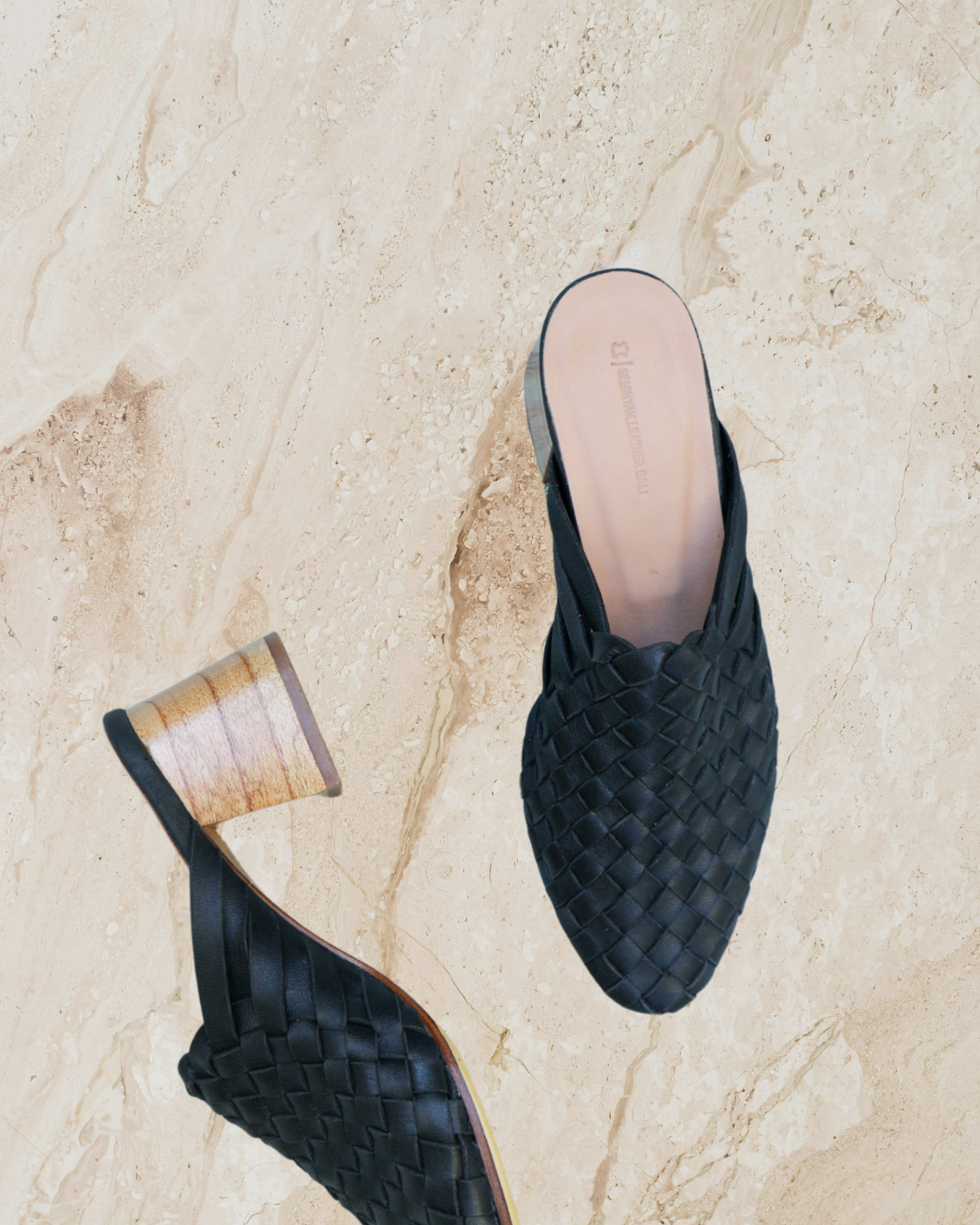 Pair of black woven mule heels designs by Seminyak Leather Bali on a beige marble surface