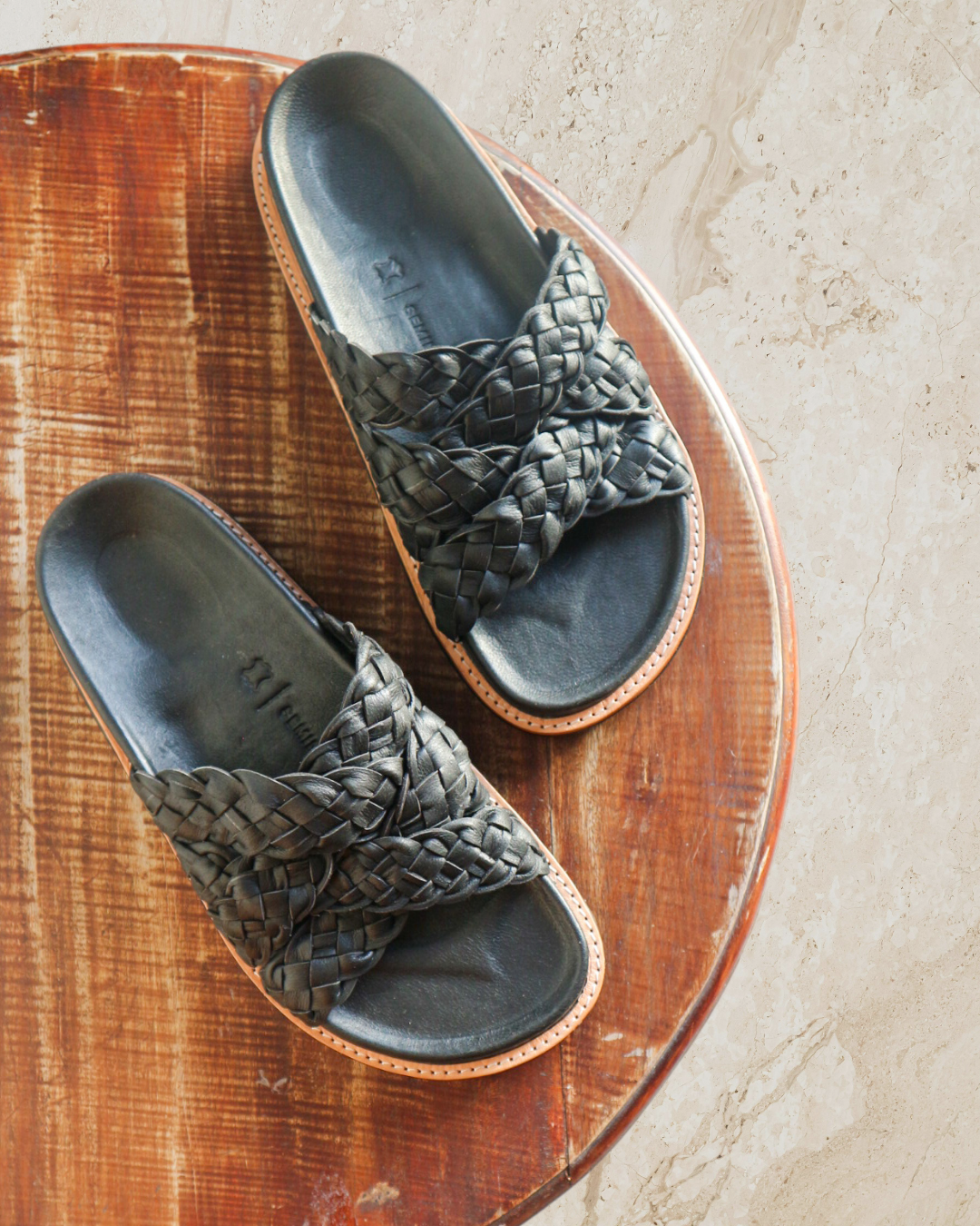 A pair of black cross woven slides with anatomic footbed displayed against a wooden table designs by Seminyak Leather Bali