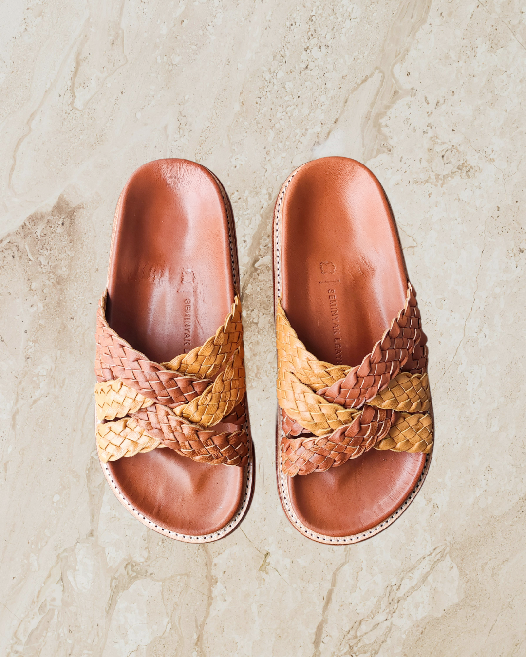 A pair of summer honey tan cross woven slides with anatomic footbed displayed against a marble background designs by Seminyak Leather Bali