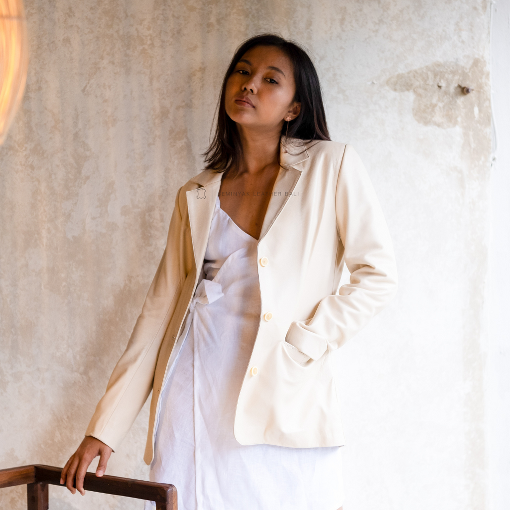 A woman wearing a beige leather blazer with a oversized cut, paired with a white dress. She is standing in a room with a textured wall designs by Seminyak Leather Bali