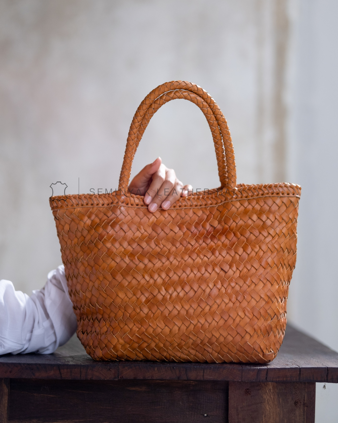 Tan woven handbag held by a person on a wooden surface with a neutral background designs by Seminya Leather Bali