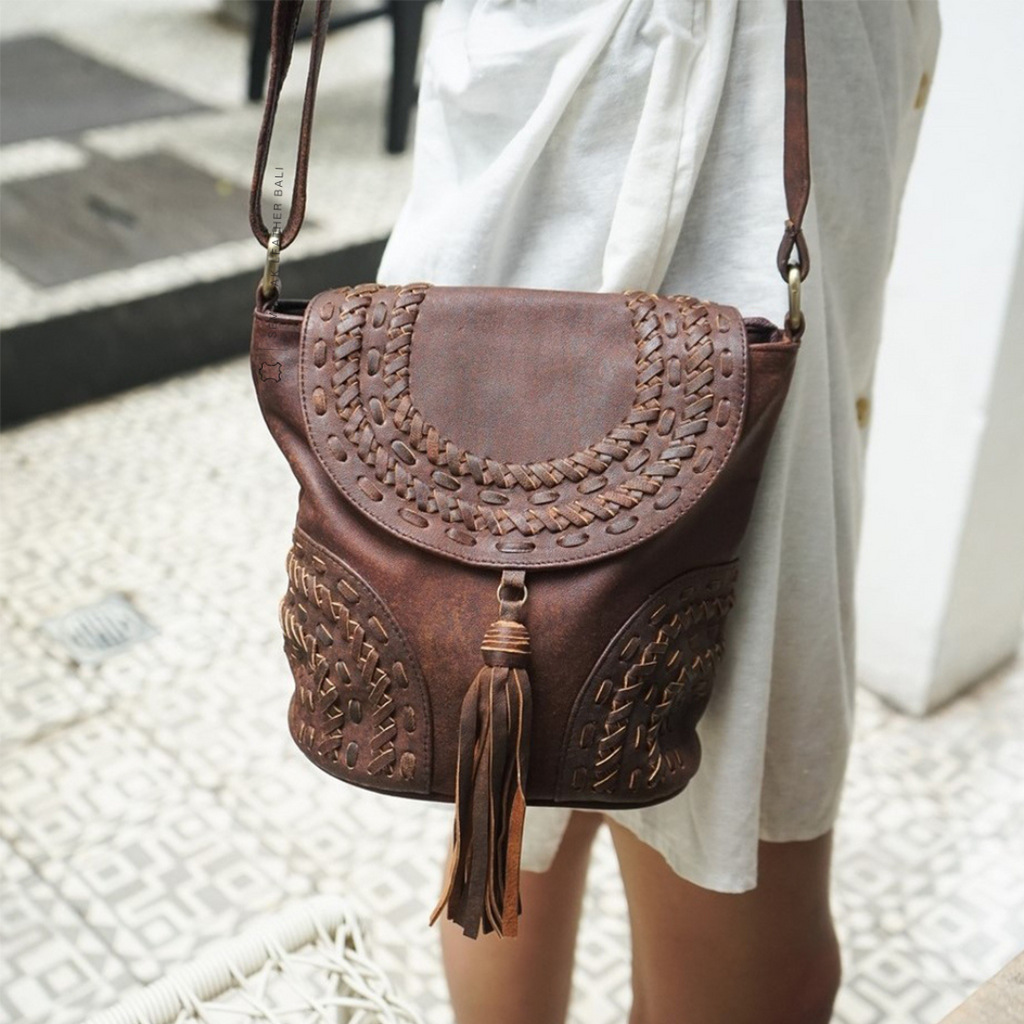 A person wearing a white dress is carrying a brown crossbody bag with woven details and a tassel designs by Seminyak Leather Bali