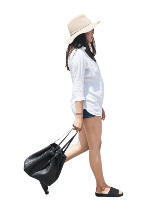 A model walks by carrying a black bag. The model wore a white shirt and jeans shorts, with additional accessories in the form of a black hat and sandals that matched the bag she was carrying. This bag is named Alice Tote Bag. The model looks comfortable carrying a bag while walking because the design of the bag is simple and comfortable to use in various ways.
