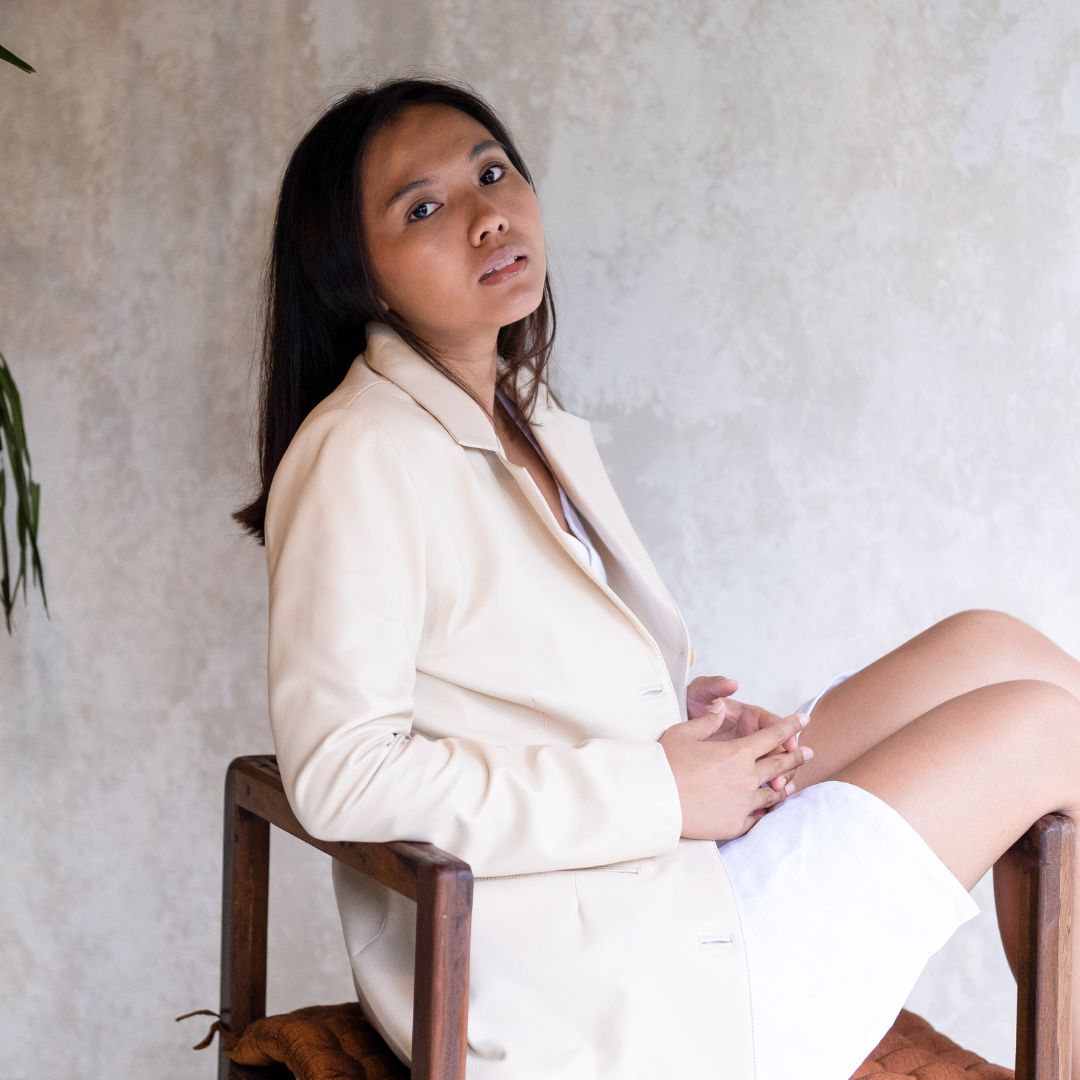 A model sits on a chair, looking up at the camera, wearing a white dress covered with a cream-colored leather jacket.