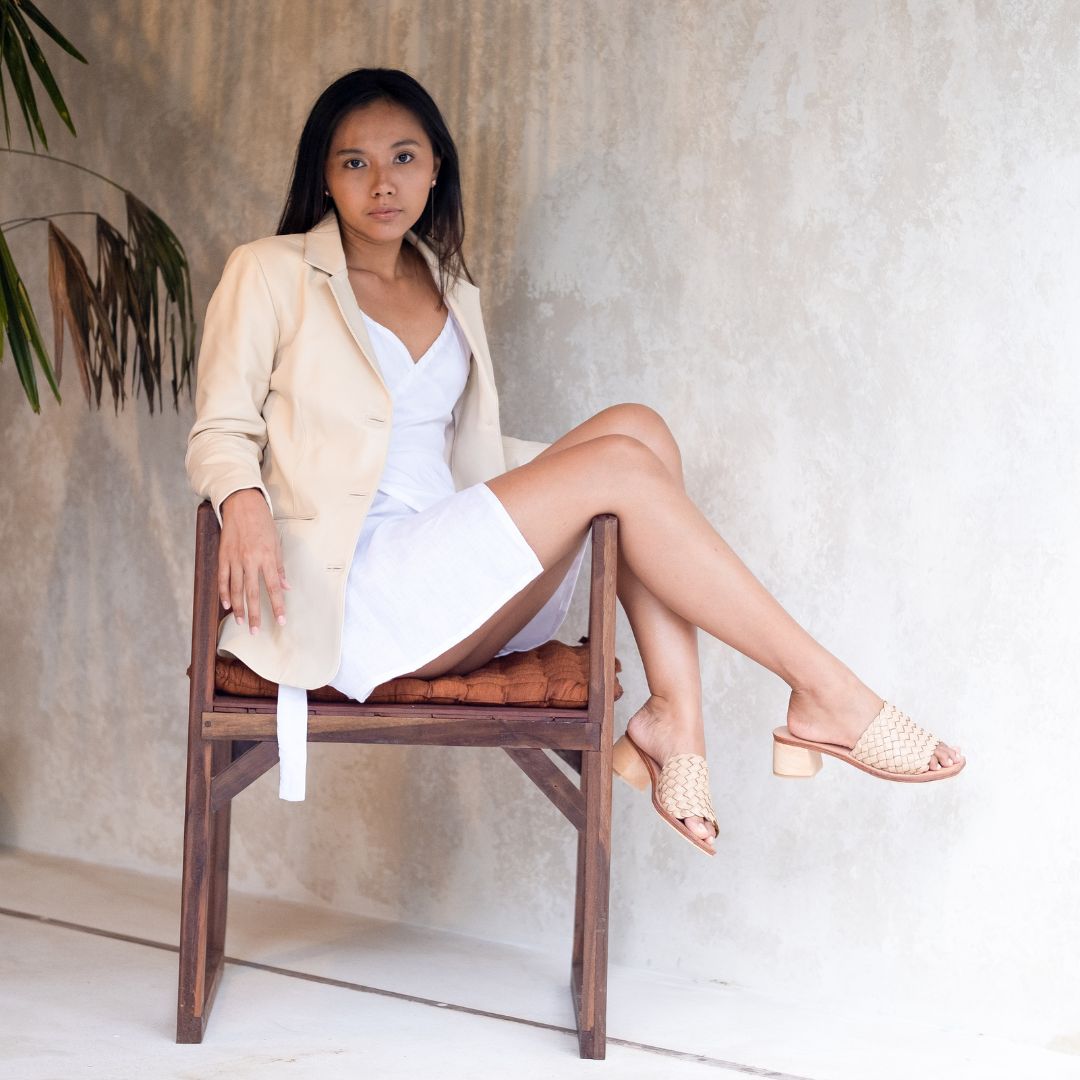 A model sits on a chair in a white dress paired with a cream-colored leather jacket. Looking straight at the camera, with one hand on the right armrest and the other on the back of the chair. Both legs are sideways and placed on the handle of the chair on the left with the addition of woven sandals with matching colors and a leather jacket that is left open when sitting