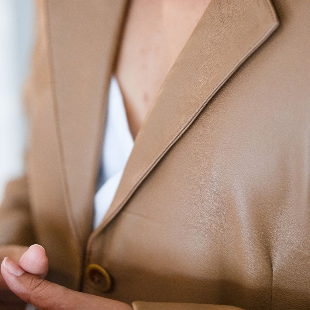 A detailed close-up of a tan leather jacket, showing the curves of the jacket's neckline and the smooth texture of the jacket. V-shaped front view of the jacket with pleat detail at the neck.
