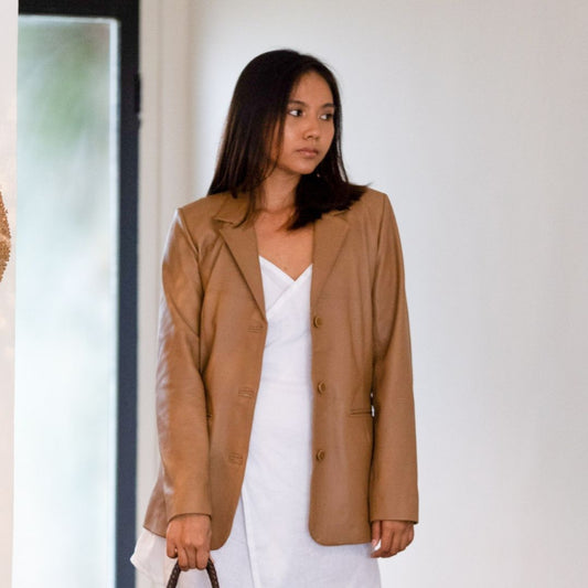 A model with shoulder-length hair stood up and glanced to the left, wearing a white dress matched with a tan leather jacket. The right hand is carrying the bag strap and the left hand is allowed to fall just like that. A leather jacket that is left open so that its function switches to outer. This jacket comes from the Seminyak Leather Bali Brand with the name Aubrey Leather Blazer