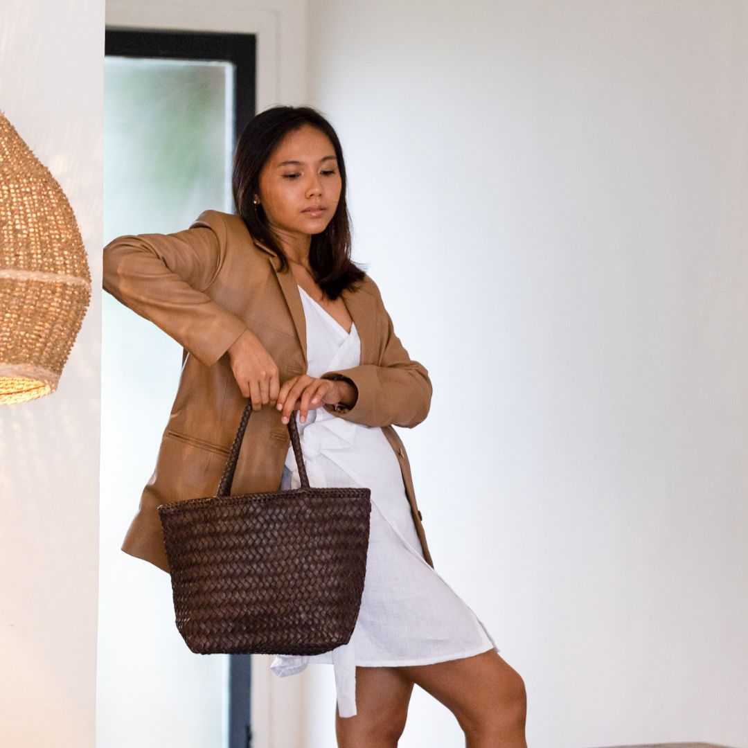 A model leaning elbows against the wall while carrying a brown handbag. By wearing a white dress suit combined with a tan leather jacket. One foot is placed in front, and the gaze is facing down. The leather jacket worn is an Aubrey Leather Blazer from the Seminyak Leather Bali brand