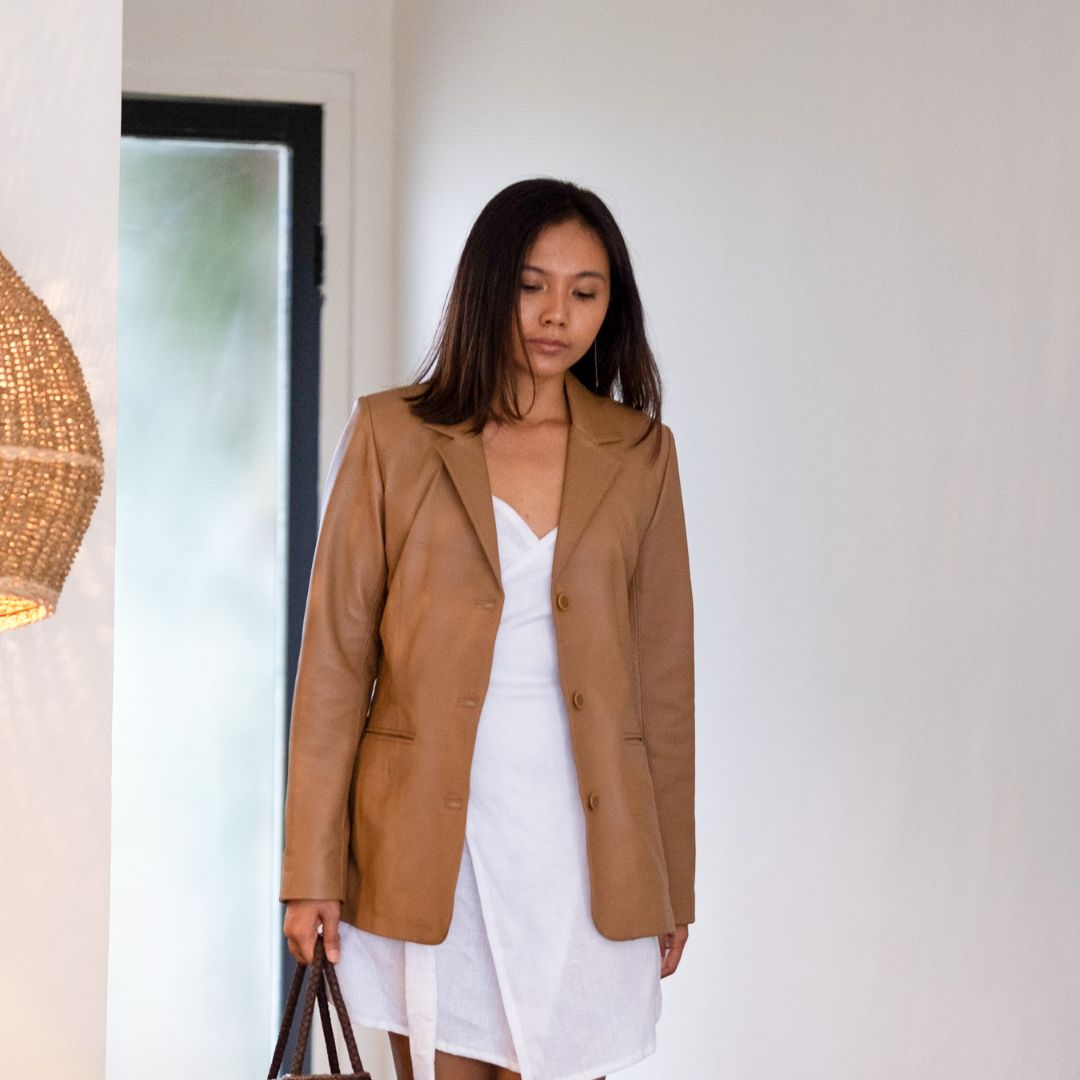 Photo of a model with shoulder-length hair walking with a downward gaze, in a white dress matched with a tan leather jacket. The jacket was left open showing the details of the white dress and carrying a brown handbag
