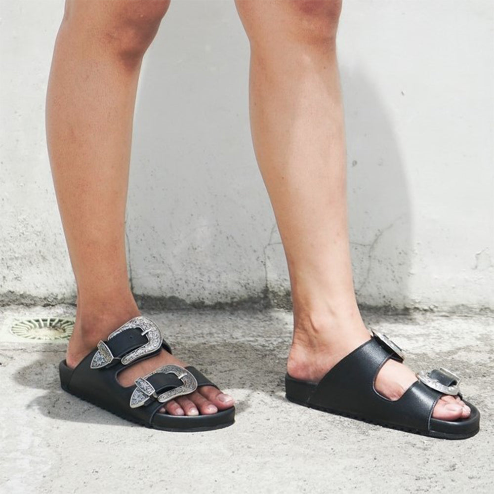 The picture shows the feet of a model wearing black sandals and a Becca crossbody bag standing. Sandals made of black cow leather, with two straps decorated with carved metal buckles, are Becky Slides Sandals from Seminyak Leather Bali
