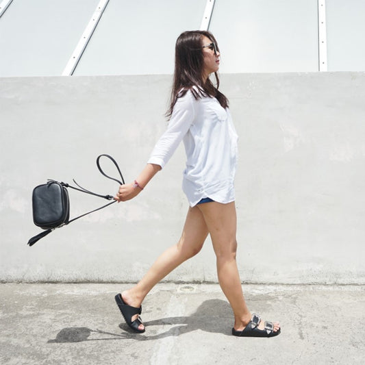 A female model walked in in a white shirt and shorts wearing black sandals and a black crossbody bag. The sandals used are made of black cowhide, which are designed to resemble slippers with two straps and are decorated with metal carving buckles. These are Becky slides Sandals from Seminyak Leather Bali