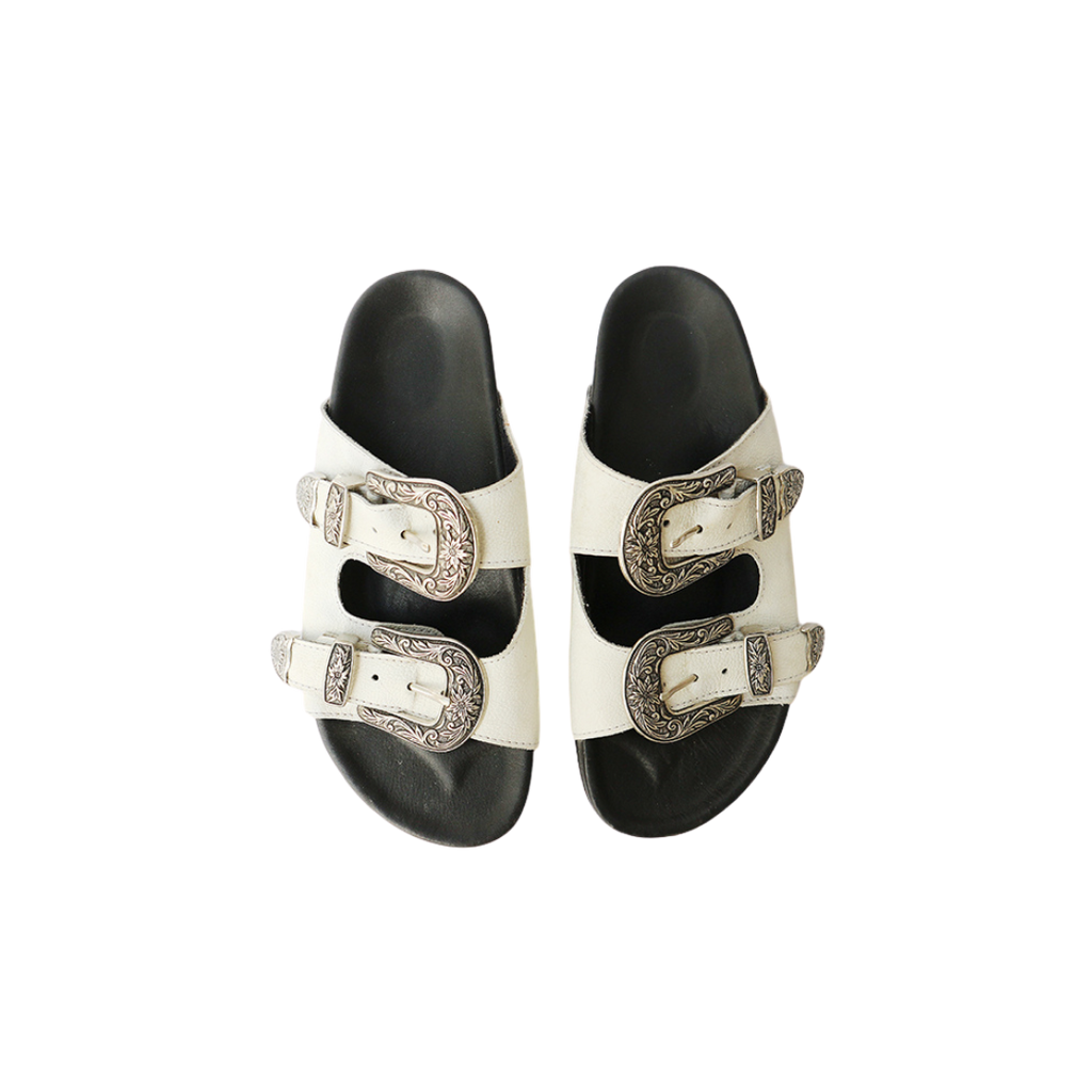 The image of a white sandal made of cowhide, designed with two straps and a carved buckle decoration is the Becky Slides Sandal from Seminyak Leather Bali
