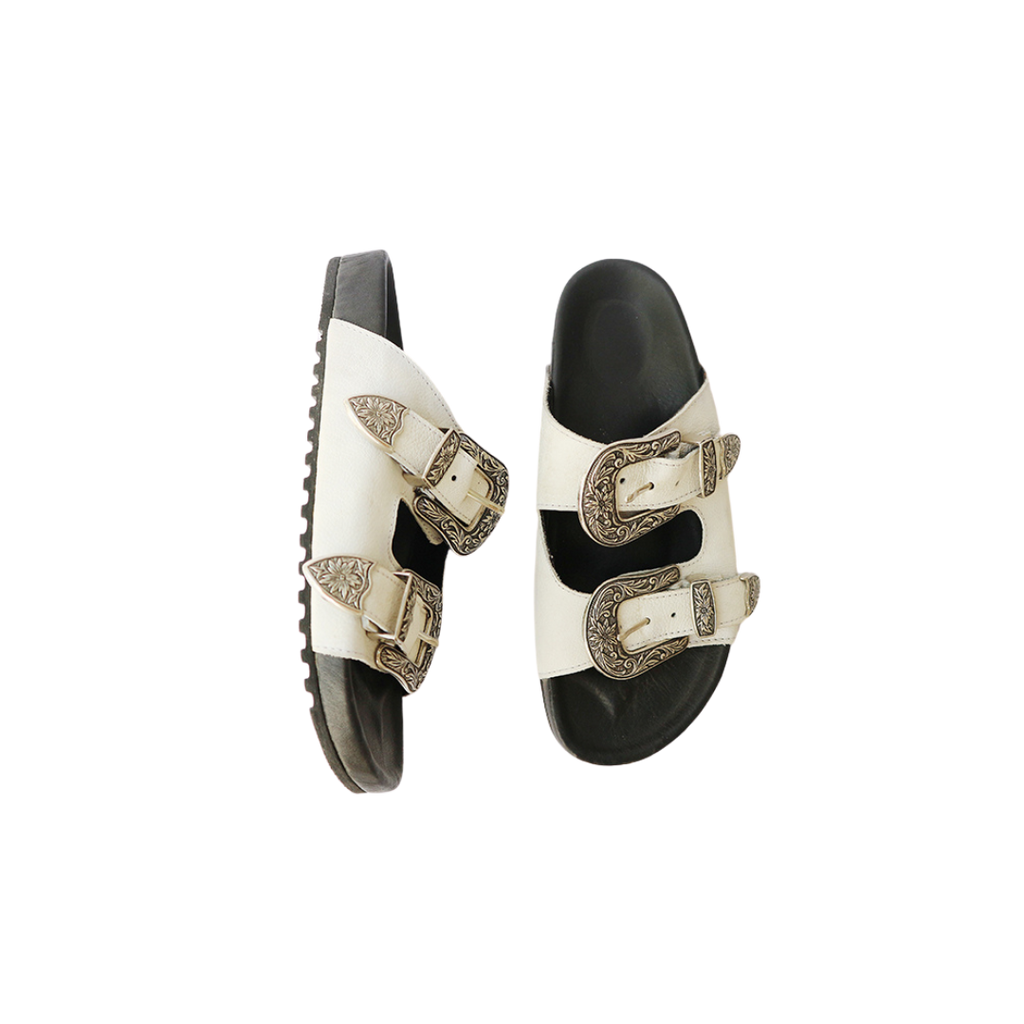 The image of a white sandal made of cowhide, designed with two straps and a carved buckle decoration is the Becky Slides Sandal from Seminyak Leather Bali