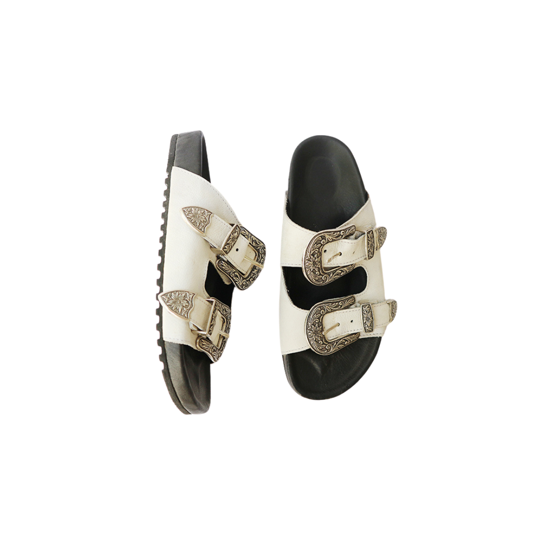The image of a white sandal made of cowhide, designed with two straps and a carved buckle decoration is the Becky Slides Sandal from Seminyak Leather Bali