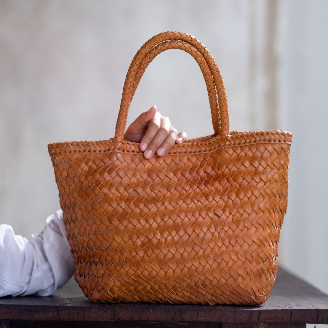 The woven bag placed on the depagang wooden table by the hands of the model is the Cening Woven Bag from Seminyak Leather Bali. This honey tan colored bag is made of vegtanned leather which is shaped by a woven method to form a strong and beautiful bag.