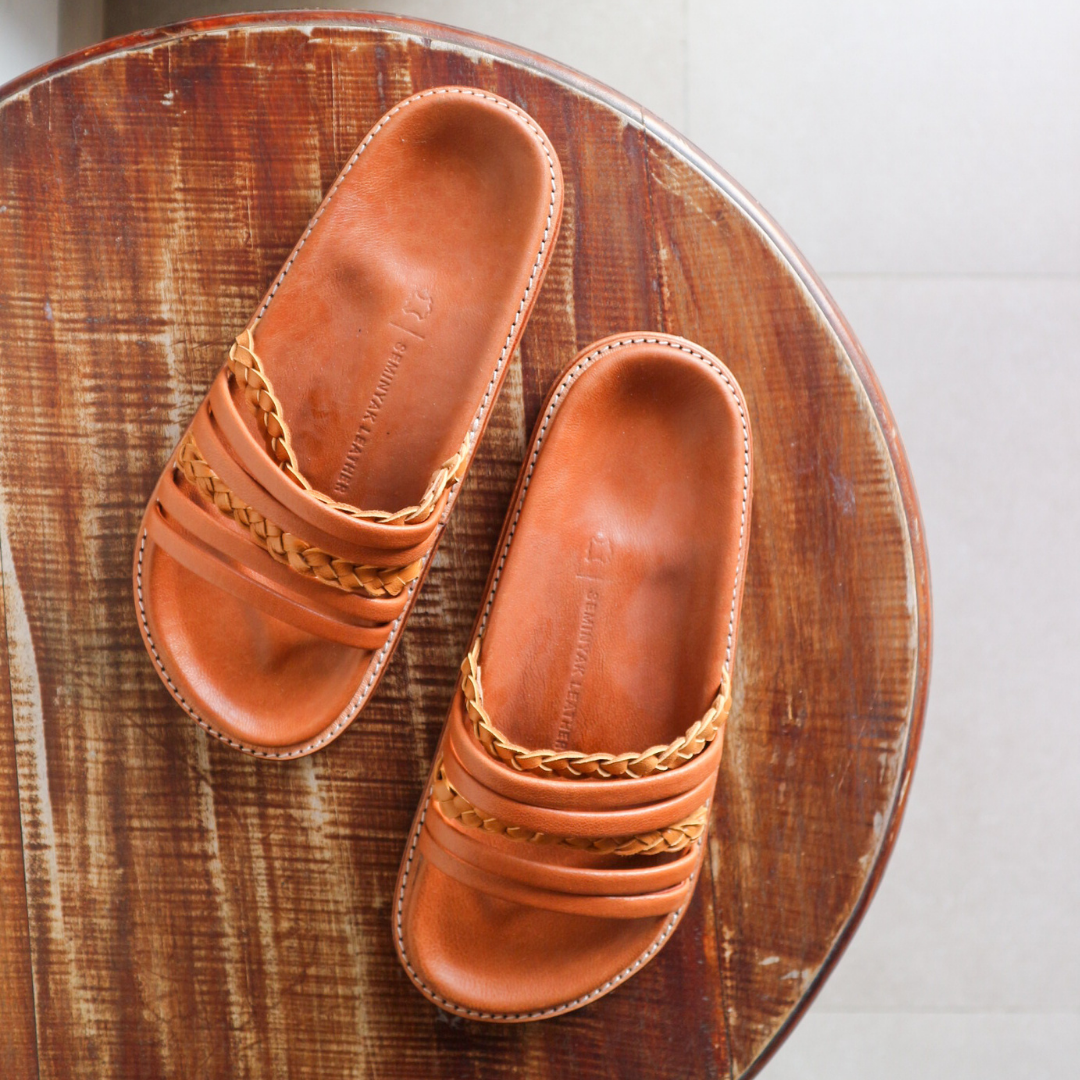 A slide is placed on the table with a color combination of summer tan and honey tan. The braid detail on the strap doesn't make the sandals look boring. These are Dayana Slides from Seminyak Leather Bali.