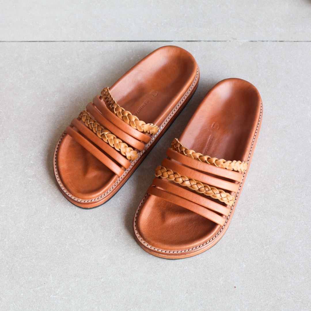 A slide is placed on the floor with a color combination of summer tan and honey tan. The braid detail on the strap doesn't make the sandals look boring. These are Dayana Slides from Seminyak Leather Bali.