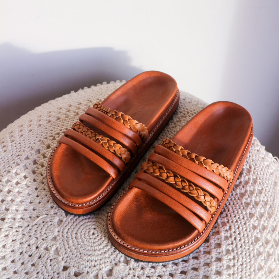 A slide is placed on the table with a color combination of summer tan and honey tan. The braid detail on the strap doesn't make the sandals look boring. These are Dayana Slides from Seminyak Leather Bali.