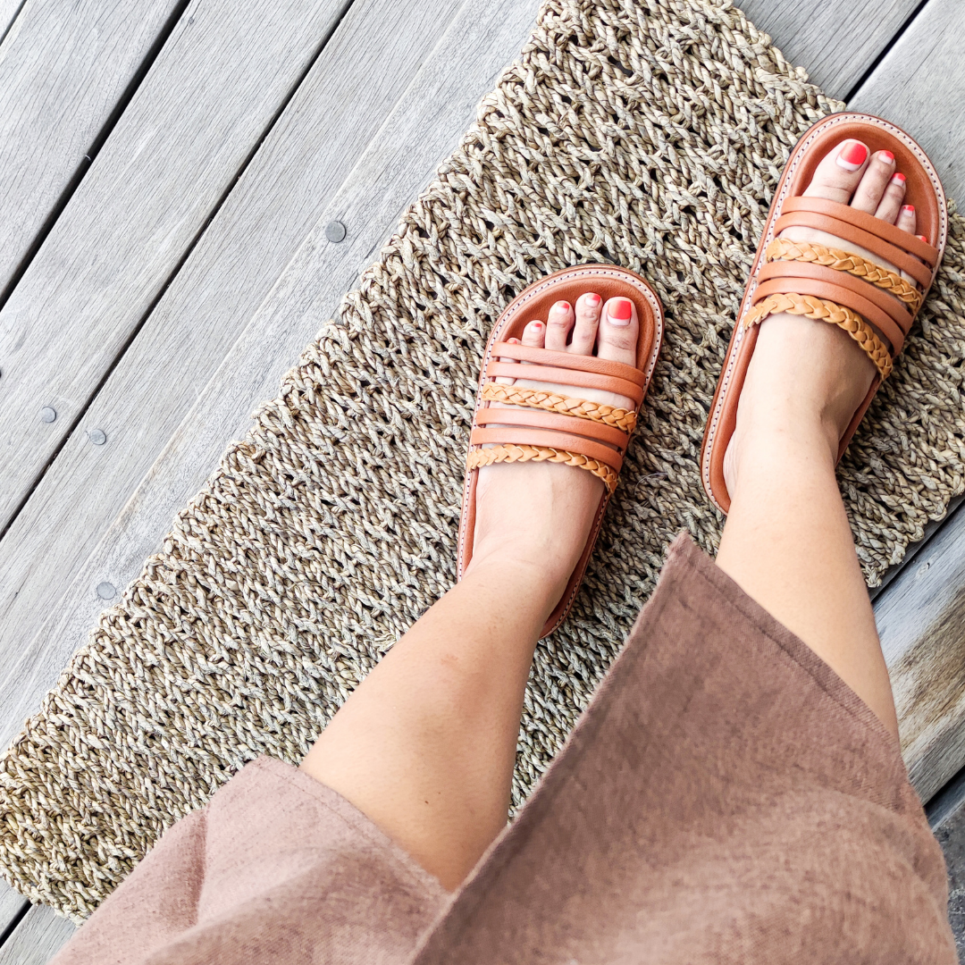 A Model shows her foot with red nails using a Sandal which have a color combination of summer tan and honey tan. The braid detail on the strap doesn't make the sandals look boring. These are Dayana Slides from Seminyak Leather Bali.