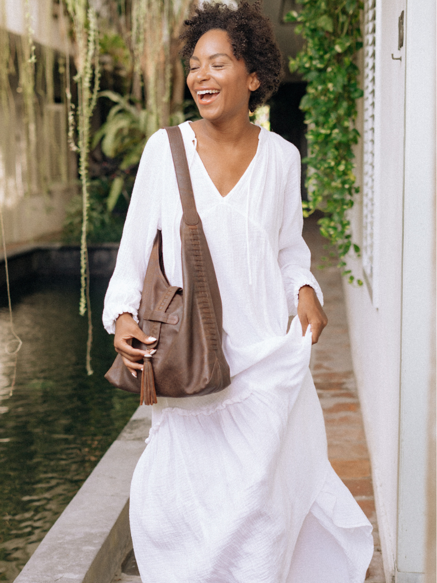 A picture shows a model with long white dress laughing and holding a beautiful hobo bag from Seminyak Leather Bali. The bag fit perfectly, so it will not prevent you from running and loughing with your friend. It is Ella Hobo Bag in Antique Brown.  
