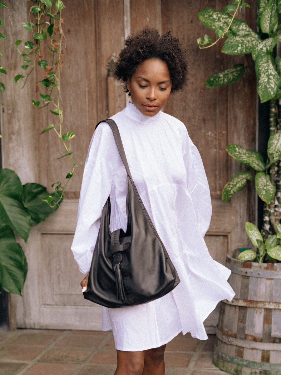 A model with white dress showing a hobo bag on her shoulder. A black tote bag, with a tassel detail for the closure, a woven detail in each side also at the front and the closure strap. This bag is Ella Hobo Bag from a magical hand of Seminyak Leather Bali