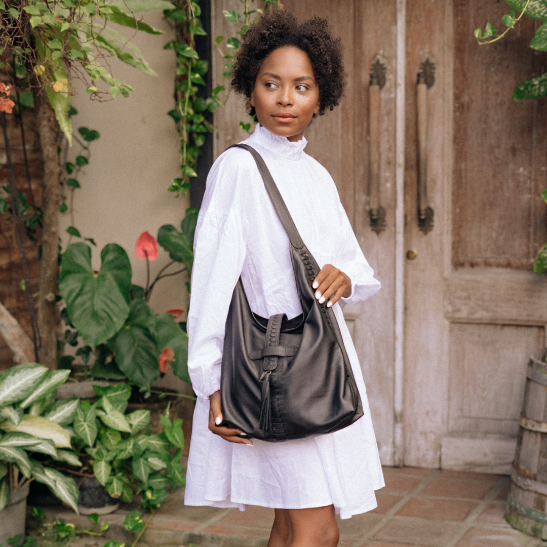 A model with white dress showing a hobo bag. A black tote bag, with a tassel detail for the closure, a woven detail in each side also at the front and the closure strap. This bag is Ella Hobo Bag from a magical hand of Seminyak Leather Bali