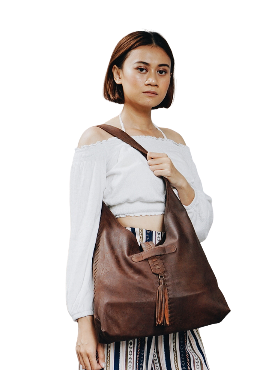 A picture shows a model with white blouse holding a bag on hel shoulder and crossed over her waist to show the detail of the bag. It is Ella Hobo Bag from Seminyak Leather Bali. 