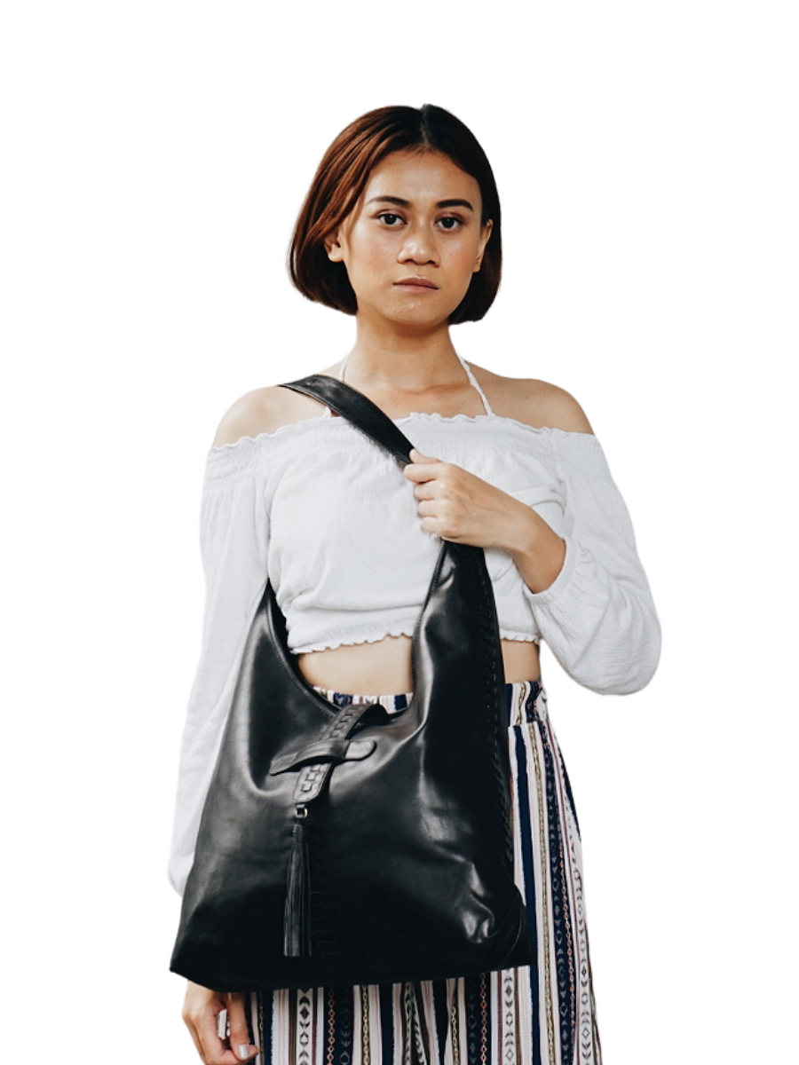 A picture shows a model with white blouse holding a bag on her shoulder and crossed over her waist to show the detail of the bag. It is Ella Hobo Bag in Black from Seminyak Leather Bali.
