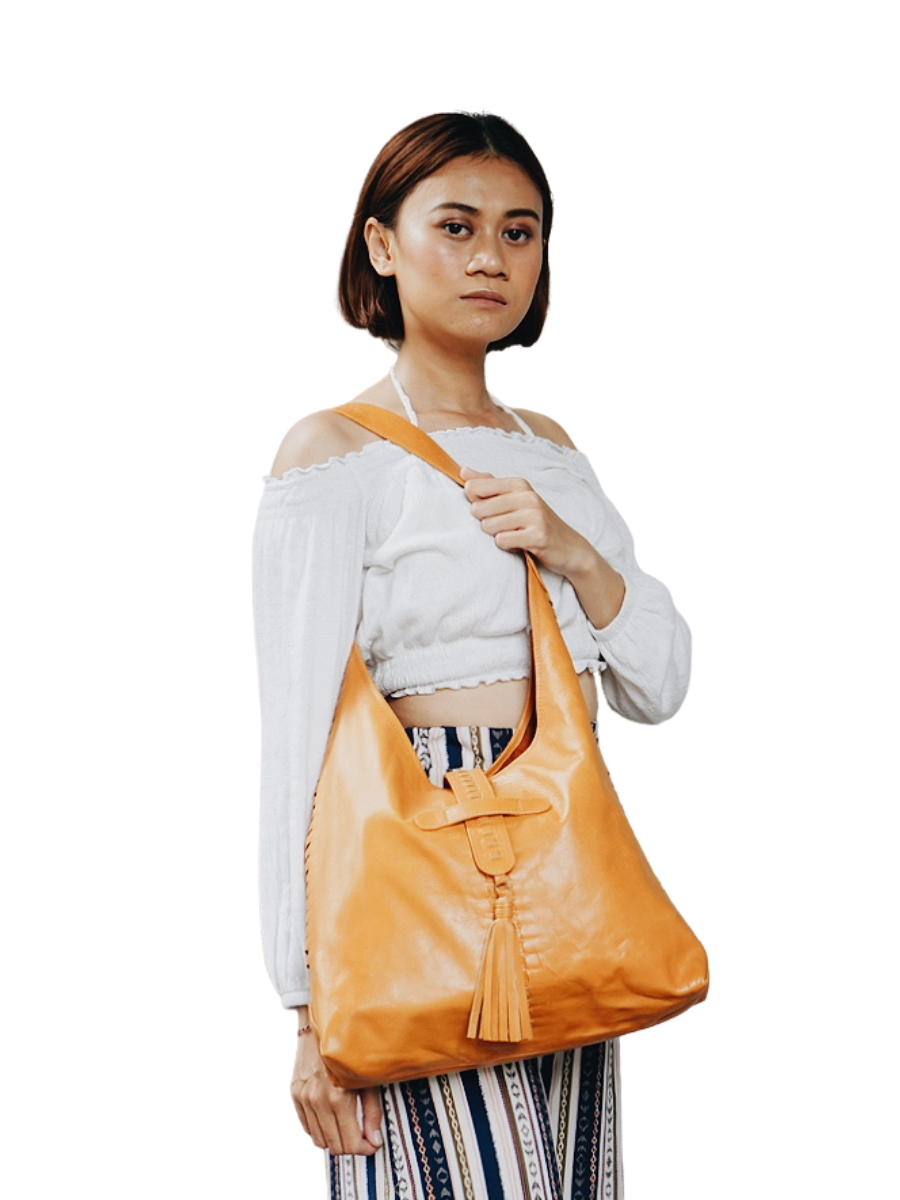 A picture shows a model with white blouse holding a bag on hel shoulder and crossed over her waist to show the detail of the bag. It is Ella Hobo Bag in honey tan from Seminyak Leather Bali.