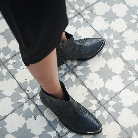 Ankle boots by Seminyak Leather Bali made of snakeskin leather in black color. Loose fit cut with carved metal detailing on the toe cap and heel. 5 cm wooden Cuban-style heel. Zipper closure. Edgy and chic footwear for bold fashion statements.
