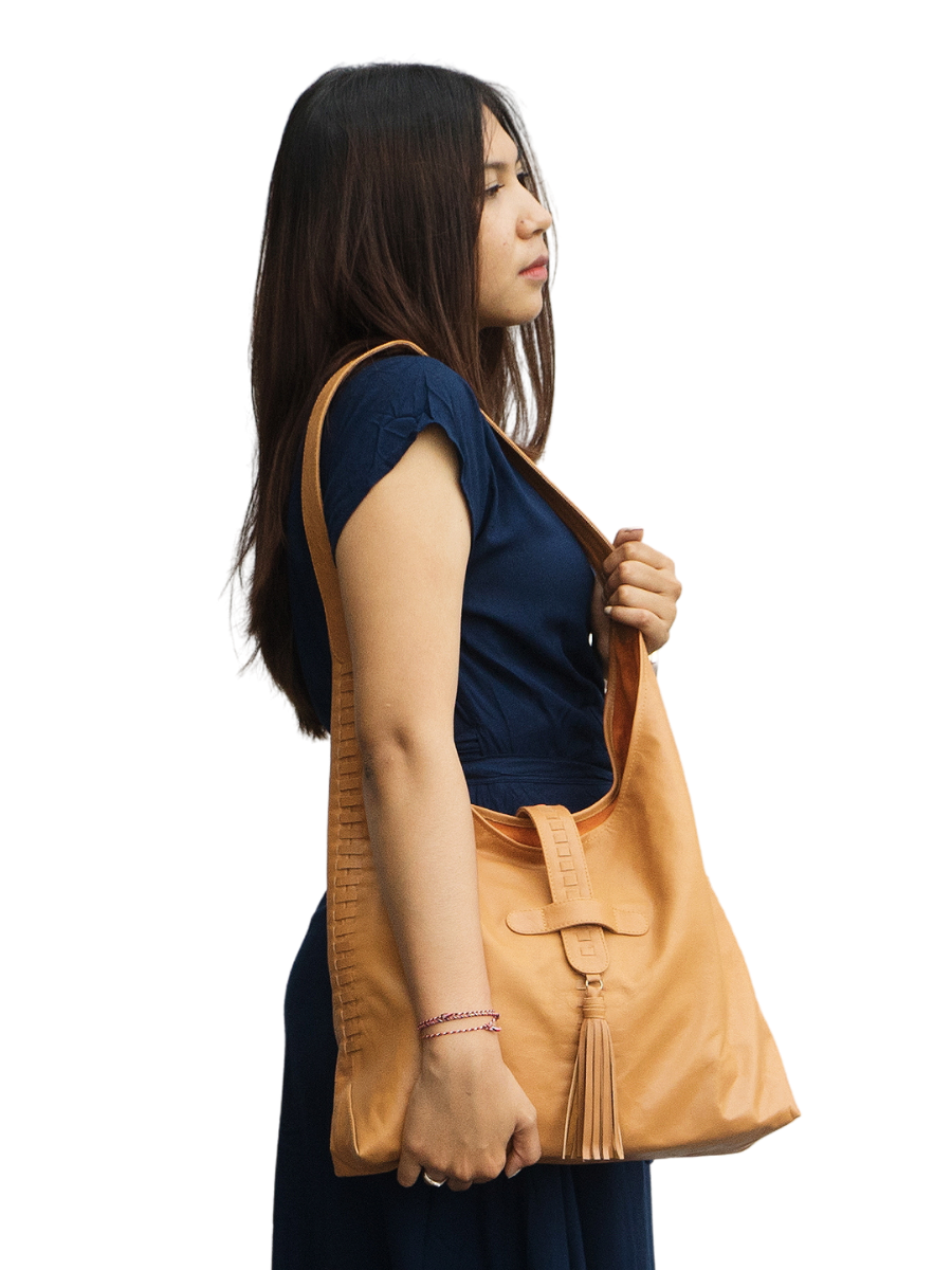 The picture shows a model with a blue dress holding a honey tan bag. The bag is made from goat leather, with a woven detail along the bag and a tassel detail in front on of the magnetic closure. The bag dimension is 41 x 31 x 10 cm. It has 1 main compartment and 1 lining pocket also a phone pocket on it. It is Ella Hobo Bag in Honey Tan from Seminyak Leather Bali.  