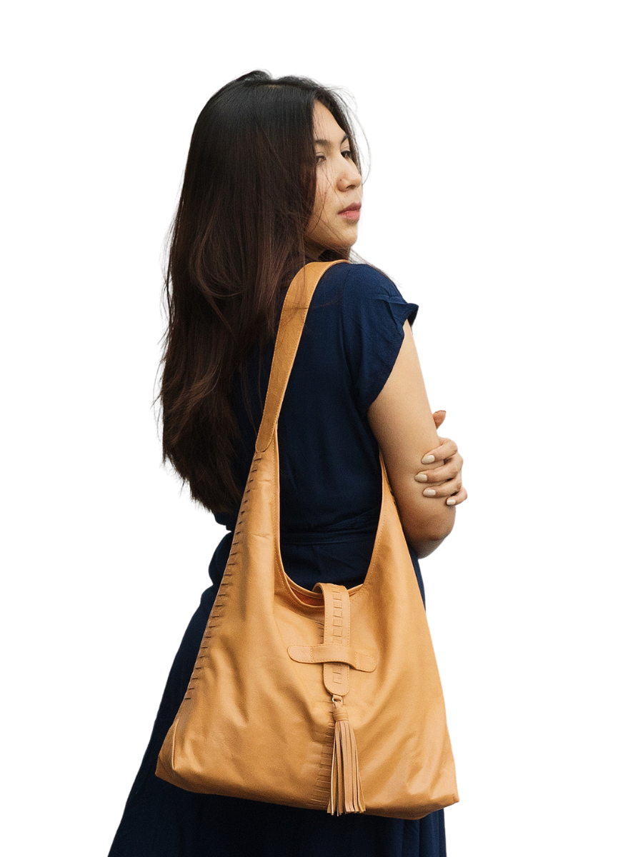 The picture shows a model with a blue dress holding a honey tan bag. The bag is made from goat leather, with a woven detail along the bag and a tassel detail in front on of the magnetic closure. The bag dimension is 41 x 31 x 10 cm. It has 1 main compartment and 1 lining pocket also a phone pocket on it. It is Ella Hobo Bag in Honey Tan from Seminyak Leather Bali.