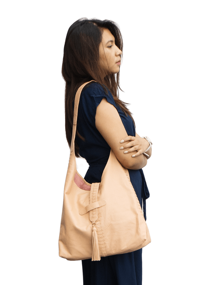 A picture shows a model in long blue dress with a bag hanging in her shoulder. The bag has a woven detail crossed in side to side and front to back, with a leather tassel in the magnetic closure. It is Ella Hobo Bag in Nude Pink from Seminyak Leather Bali.